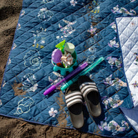 Blue quilt with beach-themed design on sand, featuring toys and sandals.