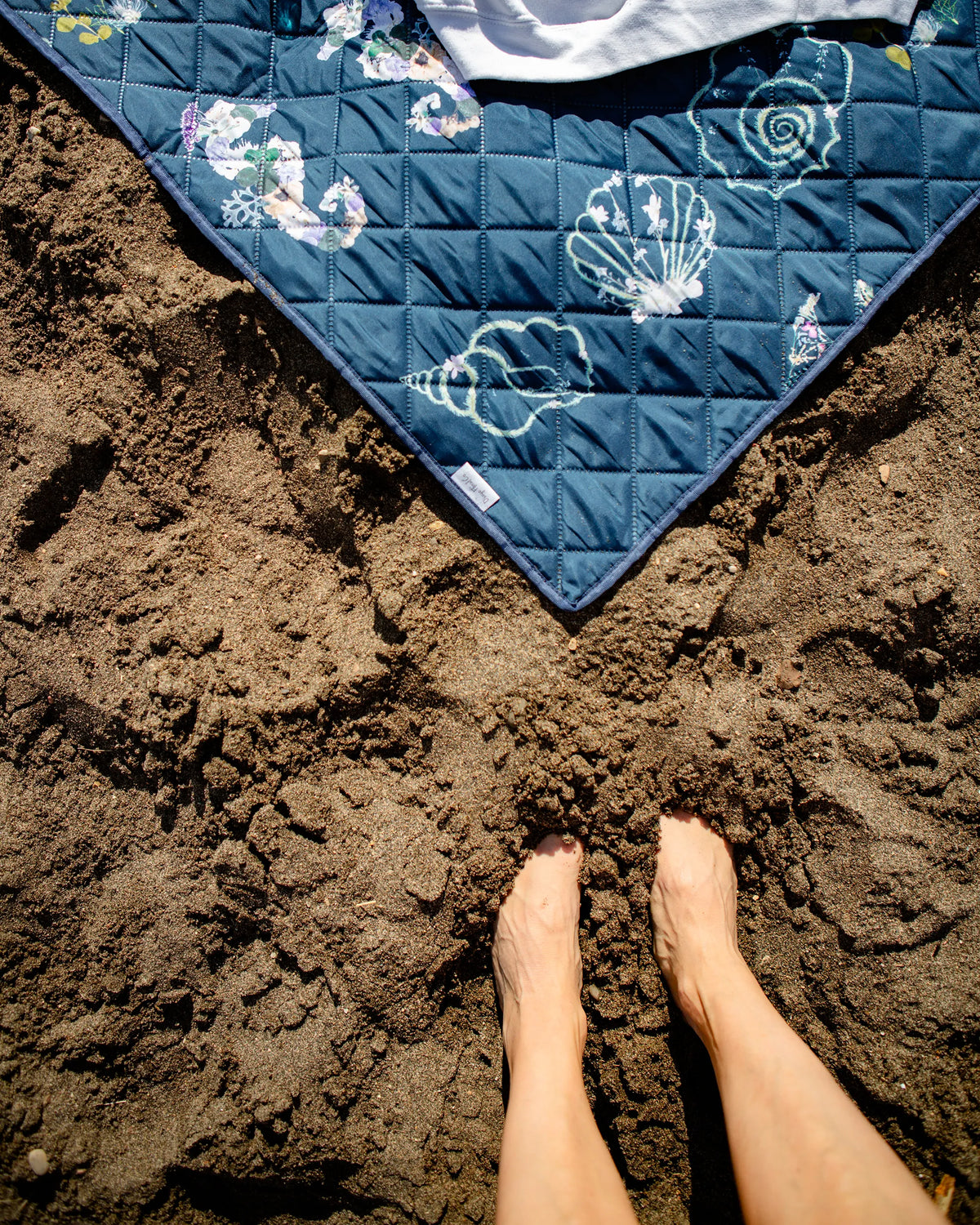 Blue outdoor blanket with shell pattern on sand, feet visible at bottom