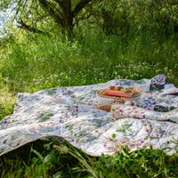 Outdoor picnic setup on a floral quilt in a grassy area with trees.