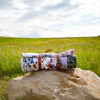 Rolled-up Oxeye Floral Co floral blanket on a rock with a grassy field and cloudy sky in the background
