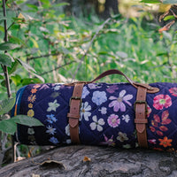 Floral-patterned picnic blanket with brown leather straps on a tree stump with green foliage in the background