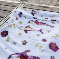 A white fabric baby blanket with a pressed flower pattern of purple whales and pressed flowers, draped on a wooden deck.