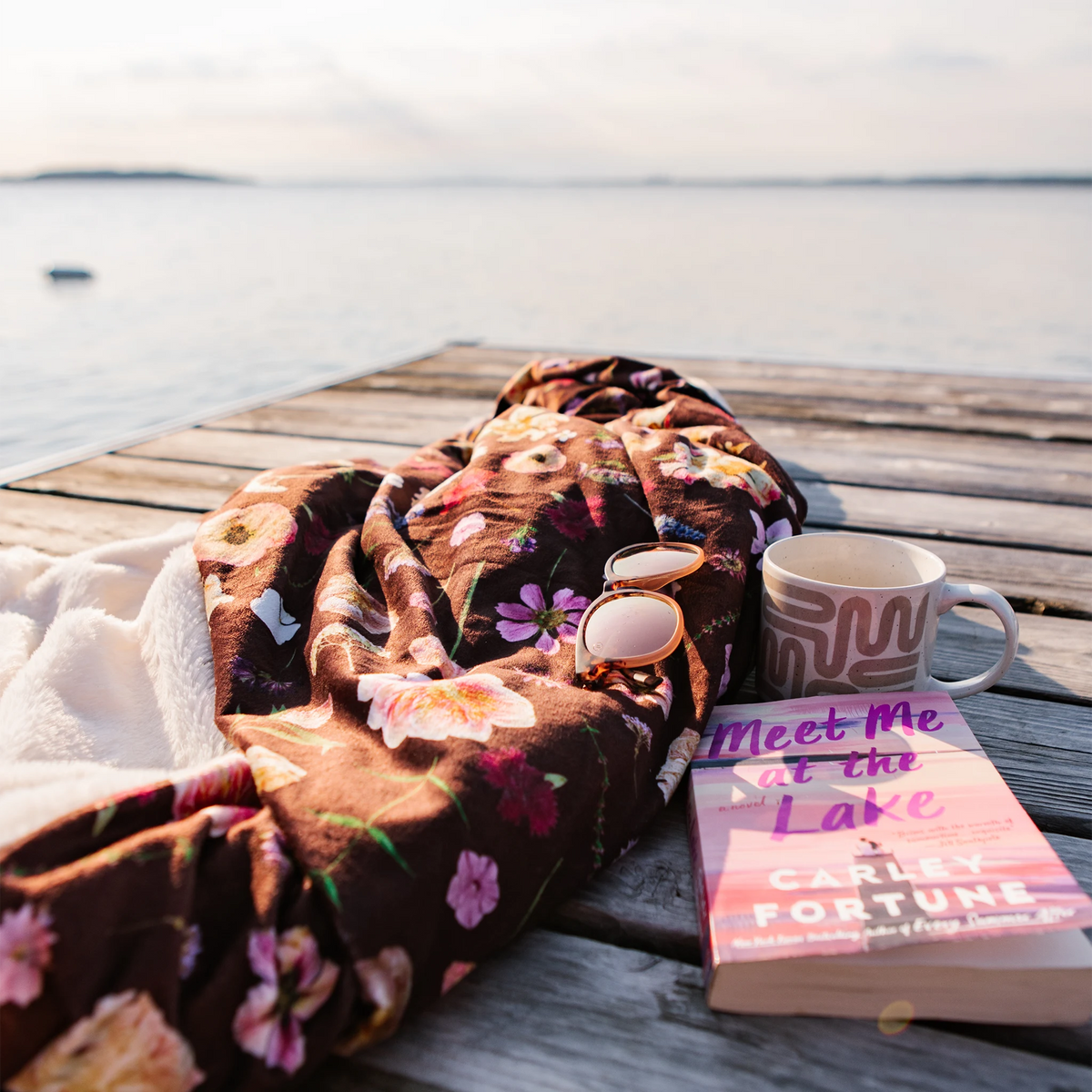 A cozy outdoor scene on a wooden dock by the water, featuring a burgundy floral blanket, a mug, sunglasses, and a pink book.