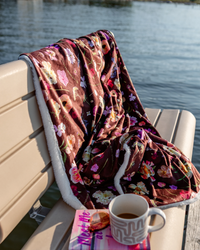 A cozy floral fleece blanket with a white sherpa lining is draped over a beige wooden dock bench next to the water. A white mug sits on a book in the foreground.