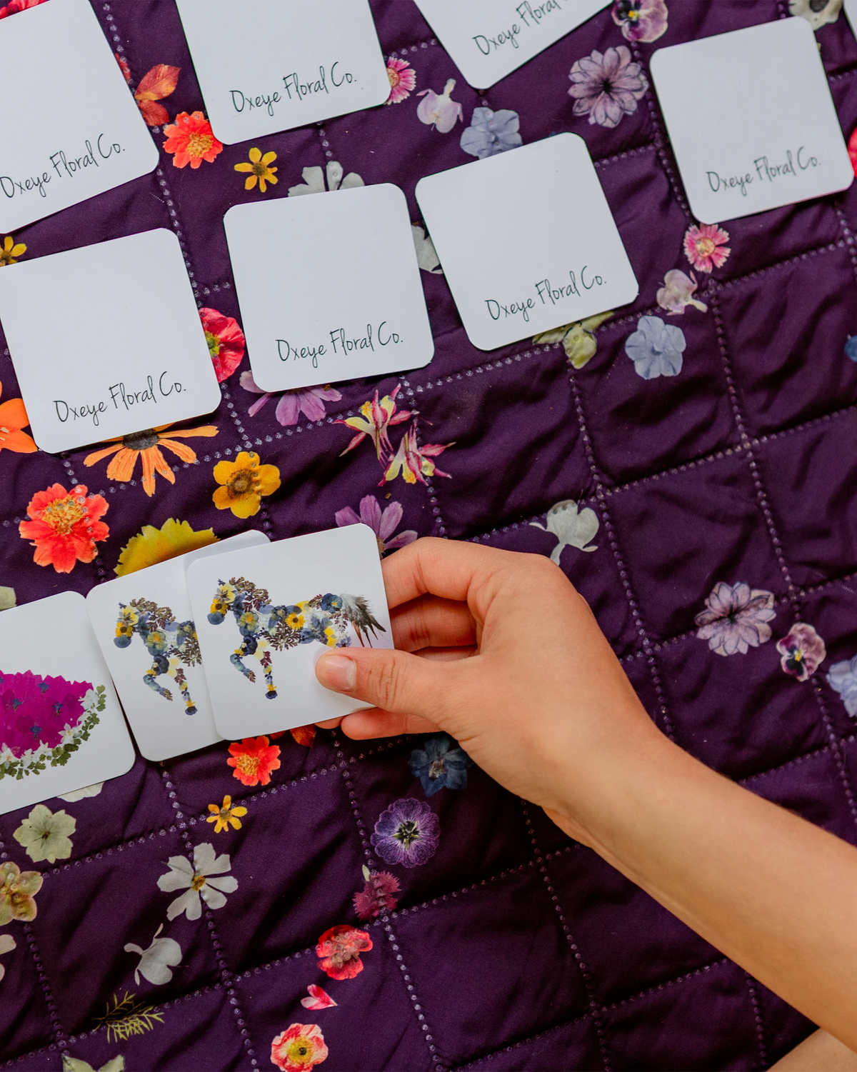 A hand is holding a small card with a colorful pressed floral horse illustration, placed over a purple quilted blanket printed with flowers. Several white cards labeled 'Oxeye Floral Co' are scattered on the quilt.