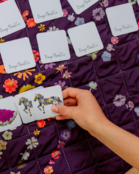 A hand is holding a small card with a colorful pressed floral horse illustration, placed over a purple quilted blanket printed with flowers. Several white cards labeled 'Oxeye Floral Co' are scattered on the quilt.