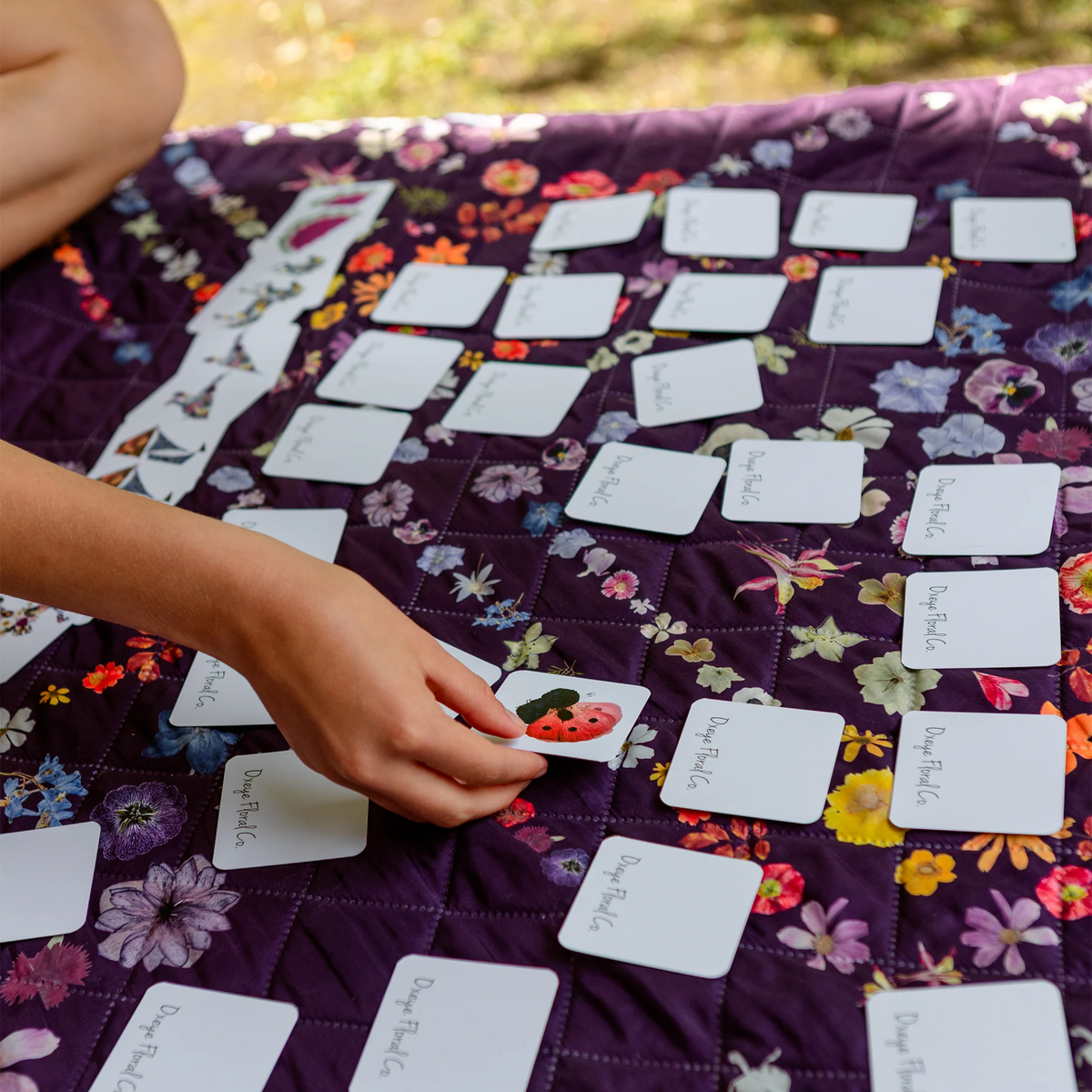 A close-up of a person's hand placing a small illustrated card onto a purple floral quilted blanket outdoors. The blanket is covered with many cards from a Memory Game set.
