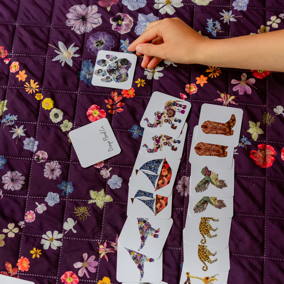 A hand is placing a memory game card on a purple quilted blanket with a floral pattern. Several cards are laid out in front, showing colorful illustrated pieces such as flowers, leaves, birds, and animals on white cardstock.