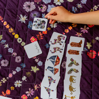 A hand is placing a memory game card on a purple quilted blanket with a floral pattern. Several cards are laid out in front, showing colorful illustrated pieces such as flowers, leaves, birds, and animals on white cardstock.