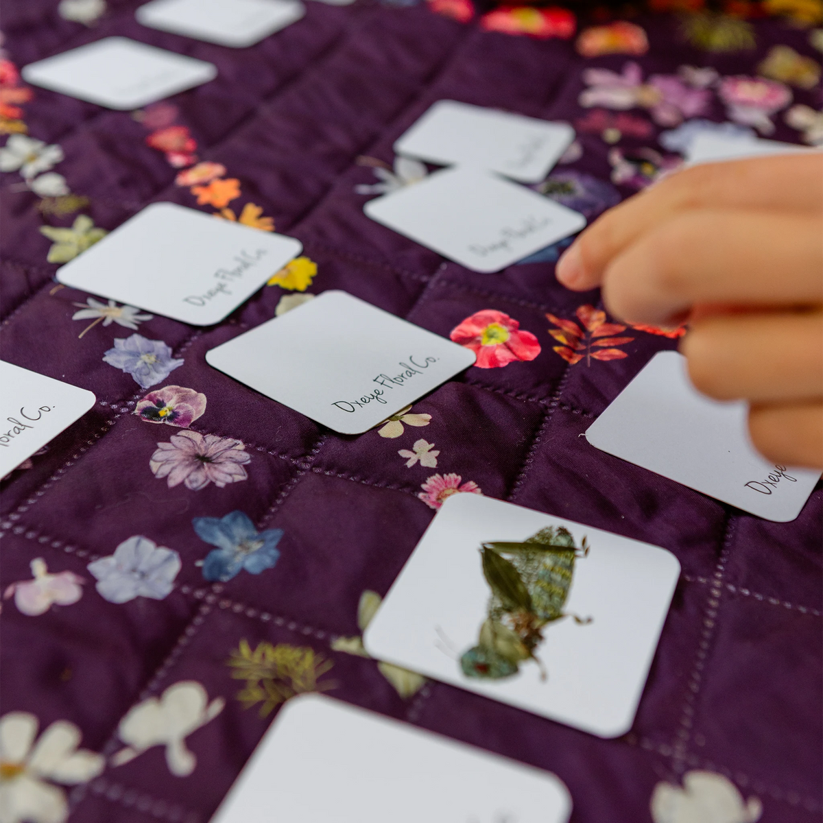 A close-up of a hand placing a white card  from a memory game onto a purple quilted fabric surface.