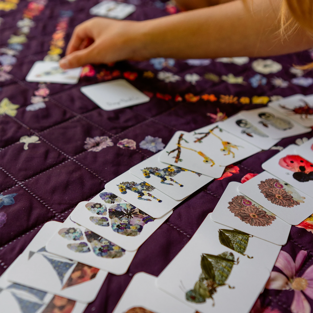 A close-up lifestyle shot of a child's hand arranging butterfly and insect cards on a purple quilted blanket with floral patterns.