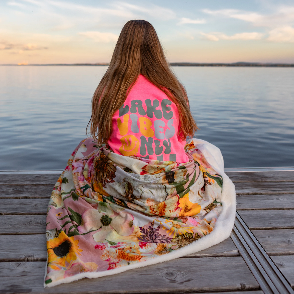 A woman sits on a wooden dock facing a calm lake at sunset, wrapped in a pressed floral blanket with a bright pink shirt that reads 'LAKE LIFE ONLY'.