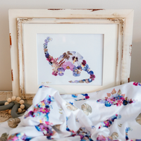 A framed floral dinosaur art print displayed in a rustic distressed white frame, styled with a matching white floral baby blanket and wooden beads on a neutral background.