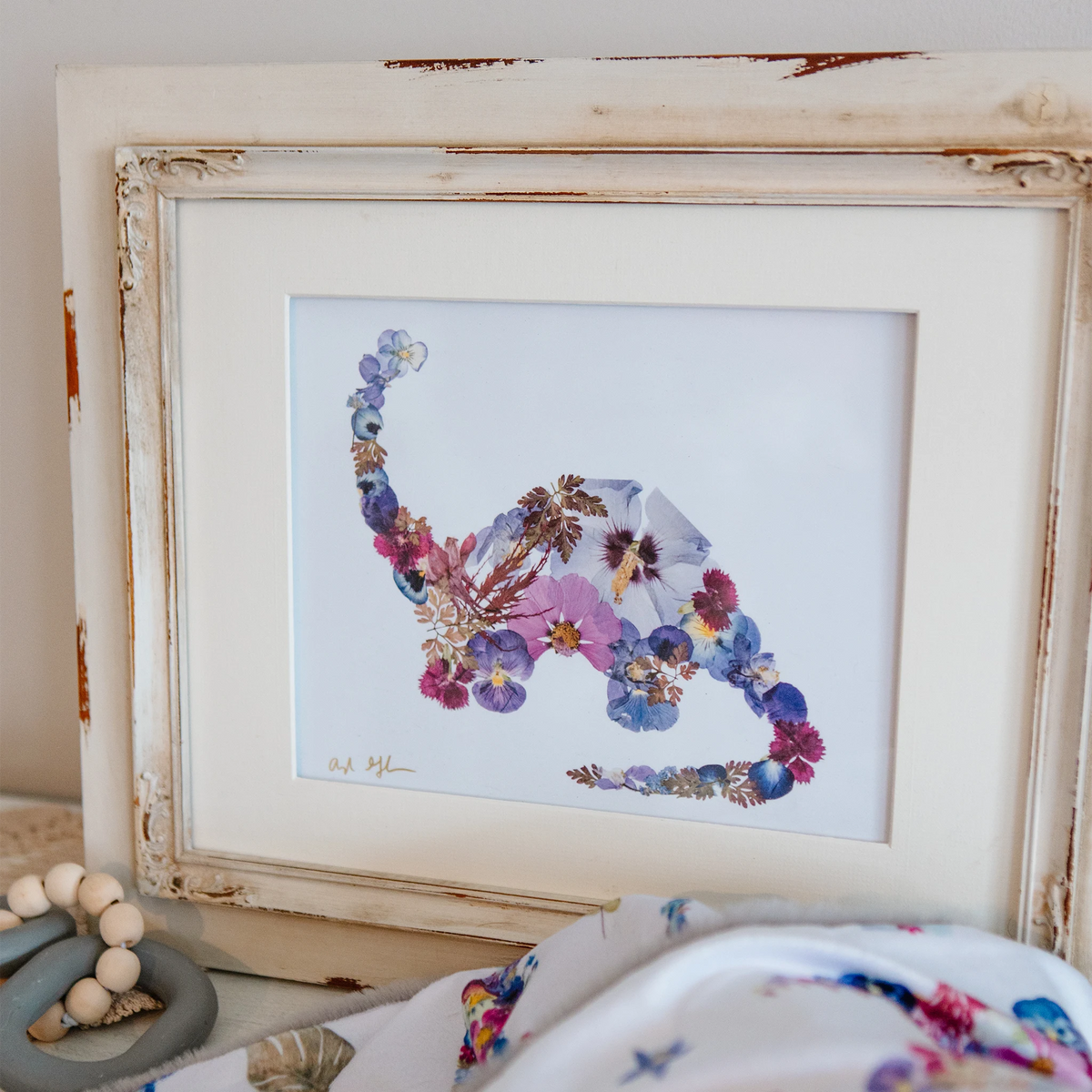 A framed floral dinosaur art print with a distressed cream-colored frame, styled on a tabletop next to a wooden bead toy and a matching floral blanket.