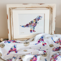 A framed floral dinosaur t-rex art print with a distressed white frame, styled on a bed with a matching white blanket featuring colorful floral tyrannosaurus patterns.