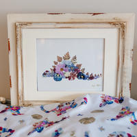 Pressed flower stegosaurus artwork in a rustic cream-coloured frame with a matching floral baby blanket in the foreground.