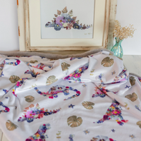 A white minky dinosaur baby blanket with a colorful floral and botanical print is draped on a surface, with a matching framed floral print and a small vase in the background.