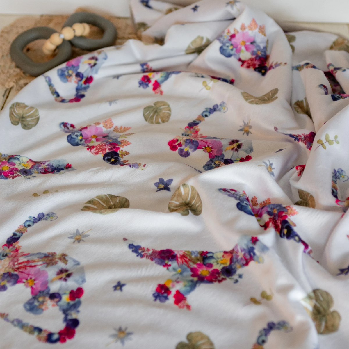 A close-up of a white baby blanket with a colorful pressed floral dinosaur print, draped in soft folds. A wooden bead teething ring is partially visible in the upper left corner.
