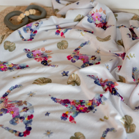 A close-up of a white baby blanket with a colorful pressed floral dinosaur print, draped in soft folds. A wooden bead teething ring is partially visible in the upper left corner.