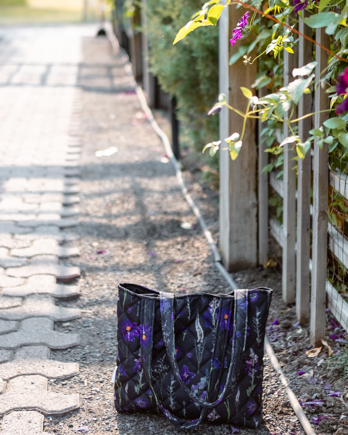 A black tote bag with a purple floral pattern sits on a paved path next to a wooden fence with green plants and purple flowers.