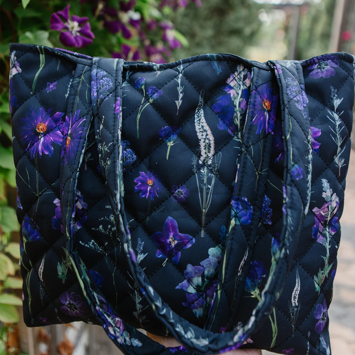 A quilted tote bag with a dark background and a purple pressed floral print, held up outdoors with blurred flowers in the background.