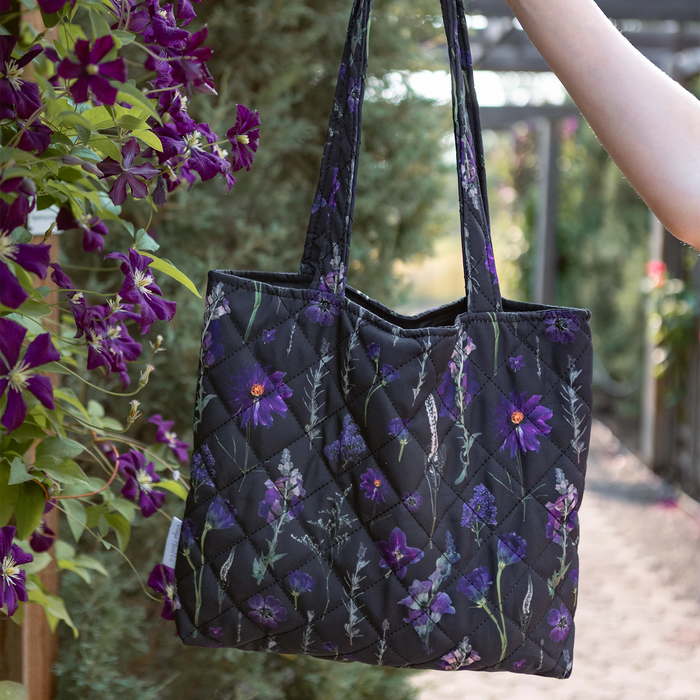 A black quilted tote bag with a purple pressed flower pattern, held by a person's arm in an outdoor garden setting with purple flowers in the foreground.