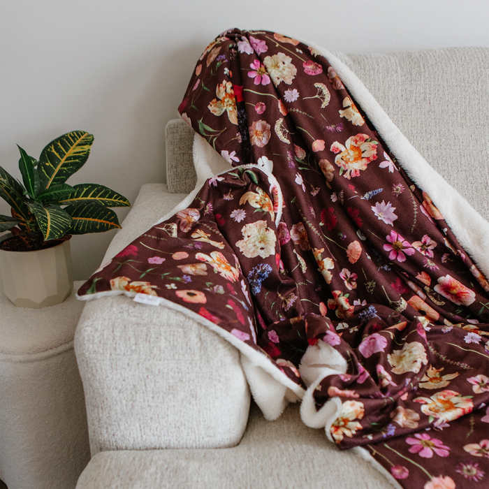 A floral throw blanket with a white sherpa fleece lining draped over a light beige armchair next to a potted green plant.