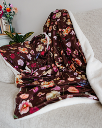A dark burgundy minky throw blanket with a colorful floral print and a white lining, draped over a light beige sofa in a living room setting.