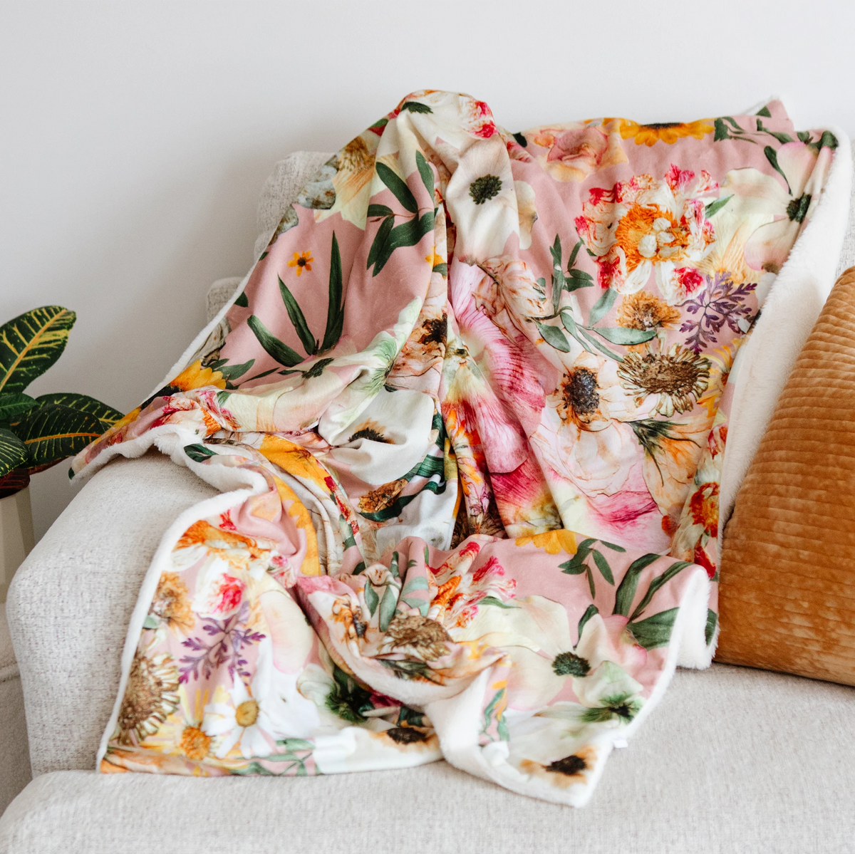 A soft floral throw blanket with a pink and cream base and green, orange, and purple botanical print, draped over a light gray upholstered armchair next to a textured mustard pillow.