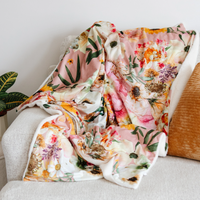 A soft floral throw blanket with a pink and cream base and green, orange, and purple botanical print, draped over a light gray upholstered armchair next to a textured mustard pillow.
