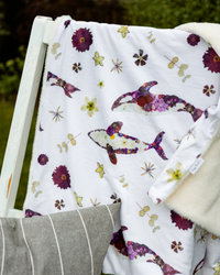 A white blanket draped over a white wooden chair, featuring a colorful pattern of purple killer whales, yellow flowers, and botanical motifs. A soft cream sherpa lining is visible on the right side, and a gray striped pillow rests at the bottom left.