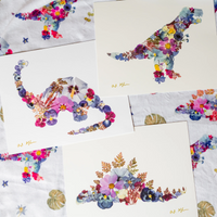 A styled flat-lay of several white art prints featuring colourful floral dinosaurs, arranged over a matching blanket.