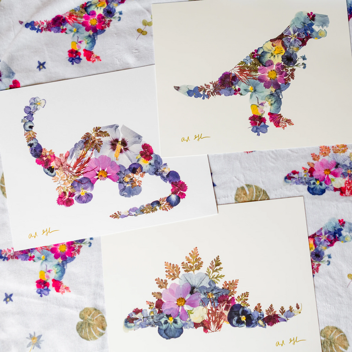 A styled flat-lay of several white art prints featuring colourful floral dinosaurs, arranged over a matching blanket.