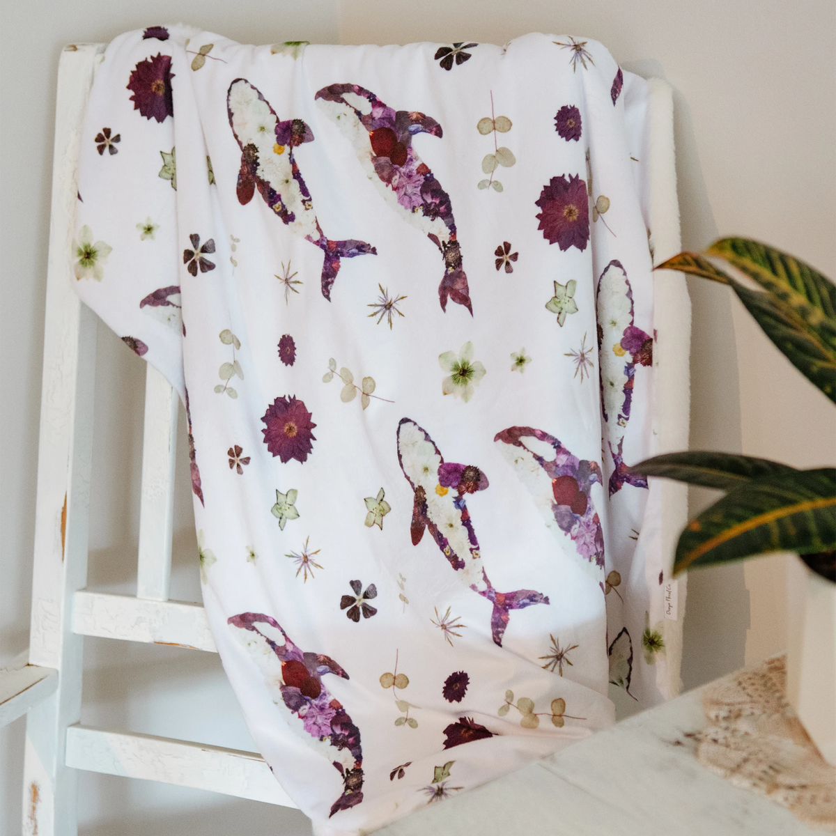 A white baby blanket with a pattern of purple orca whales and dried flowers, draped over a white wooden chair in a home setting.