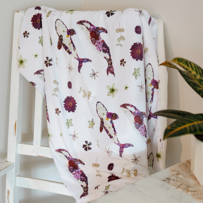 A white baby blanket with a pattern of purple orca whales and dried flowers, draped over a white wooden chair in a home setting.