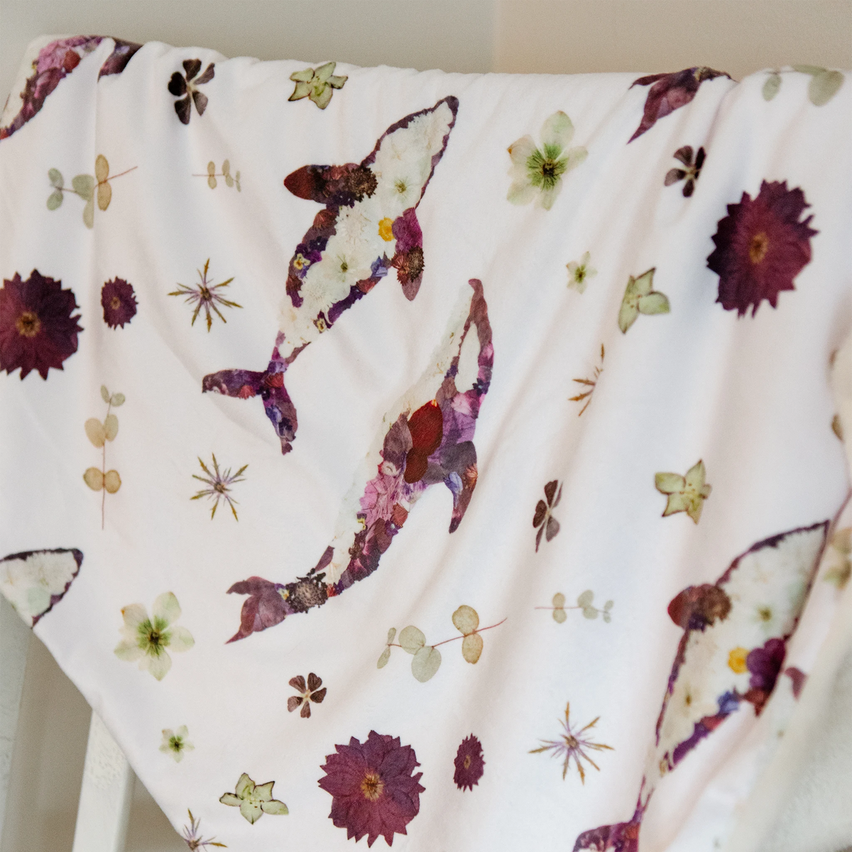 A white baby blanket draped over a white chair, featuring a pattern of pressed flowers and whales in muted purple, burgundy, and beige tones.