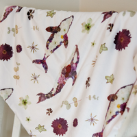 A white baby blanket draped over a white chair, featuring a pattern of pressed flowers and whales in muted purple, burgundy, and beige tones.