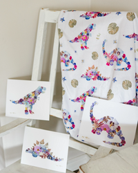 A dinosaur baby blanket and matching art prints featuring floral dinosaur pictures made of colorful flowers, displayed on a white wooden chair.