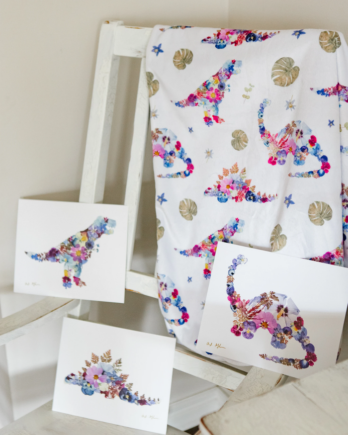 A dinosaur baby blanket and matching art prints featuring floral dinosaur pictures made of colorful flowers, displayed on a white wooden chair.