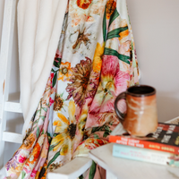 Oxeye Floral Co Peaches and Sunshine Throw Blanket draped on a chair with a coffee cup and books in the foreground.