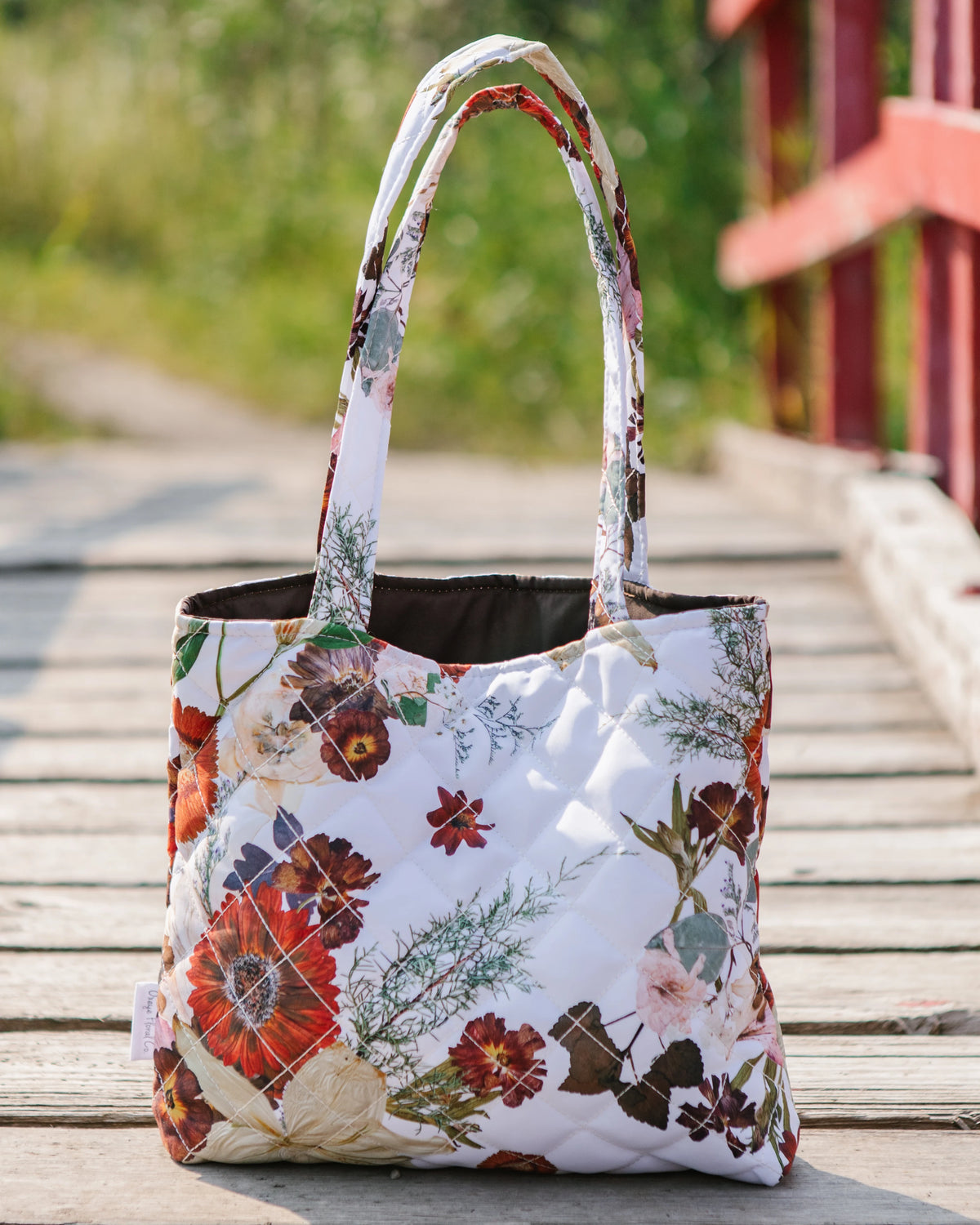 A quilted tote bag with a white background and a colorful floral print, featuring two shoulder straps, set on a wooden deck with a blurred outdoor background.