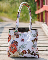 A quilted tote bag with a white background and a colorful floral print, featuring two shoulder straps, set on a wooden deck with a blurred outdoor background.
