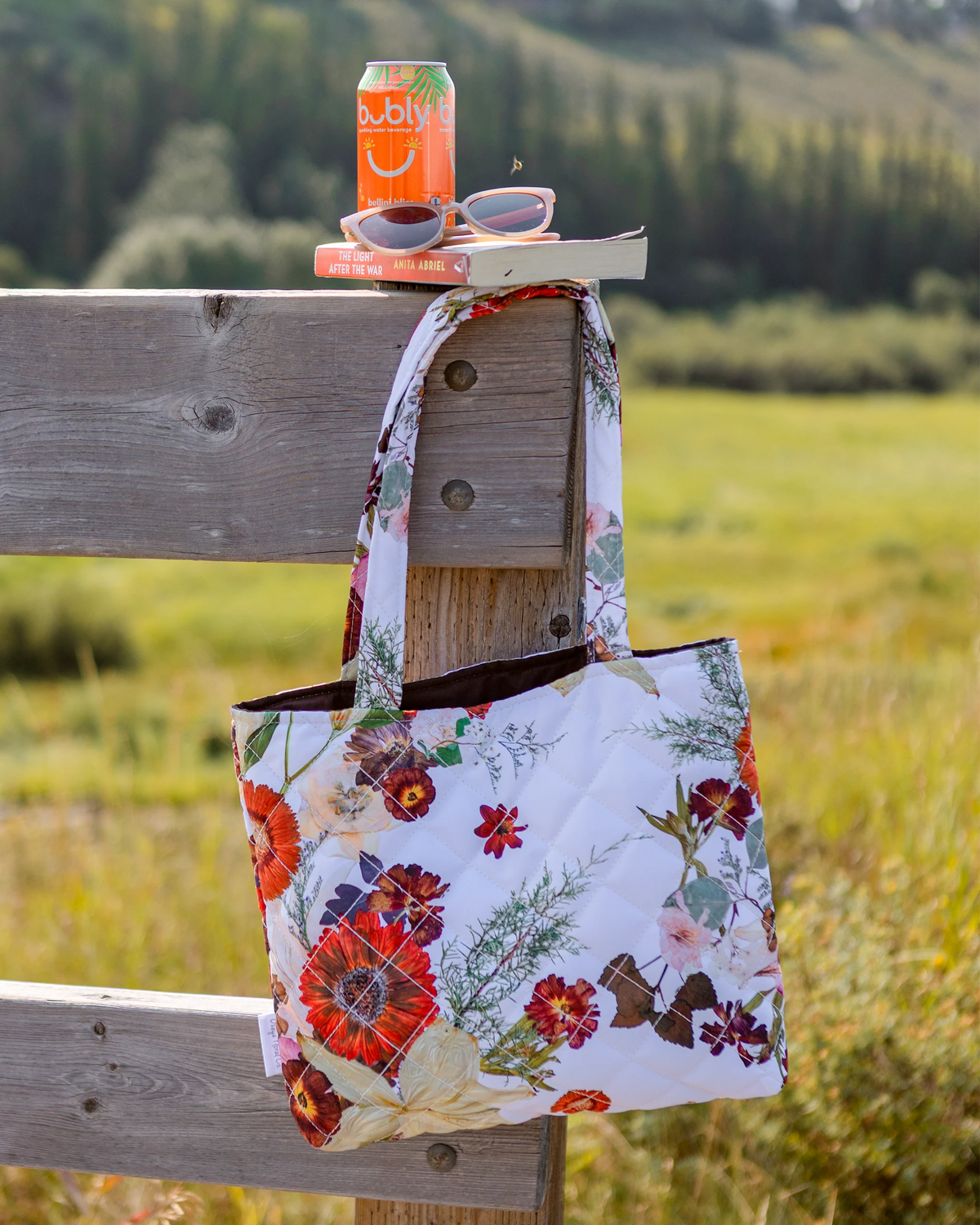 Pressed flower tote hanging on a rustic wooden fence with soda can, sunglasses and book on top of the post.