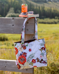 Pressed flower tote hanging on a rustic wooden fence with soda can, sunglasses and book on top of the post.