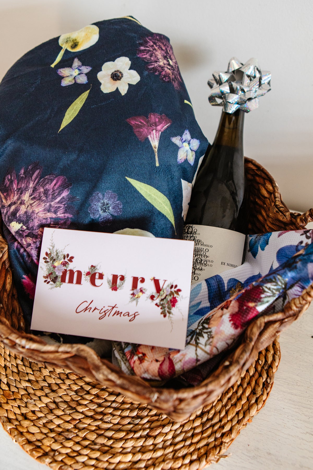 Bottle of champagne with floral blanket in a gift basket and a 'Merry Christmas' card.