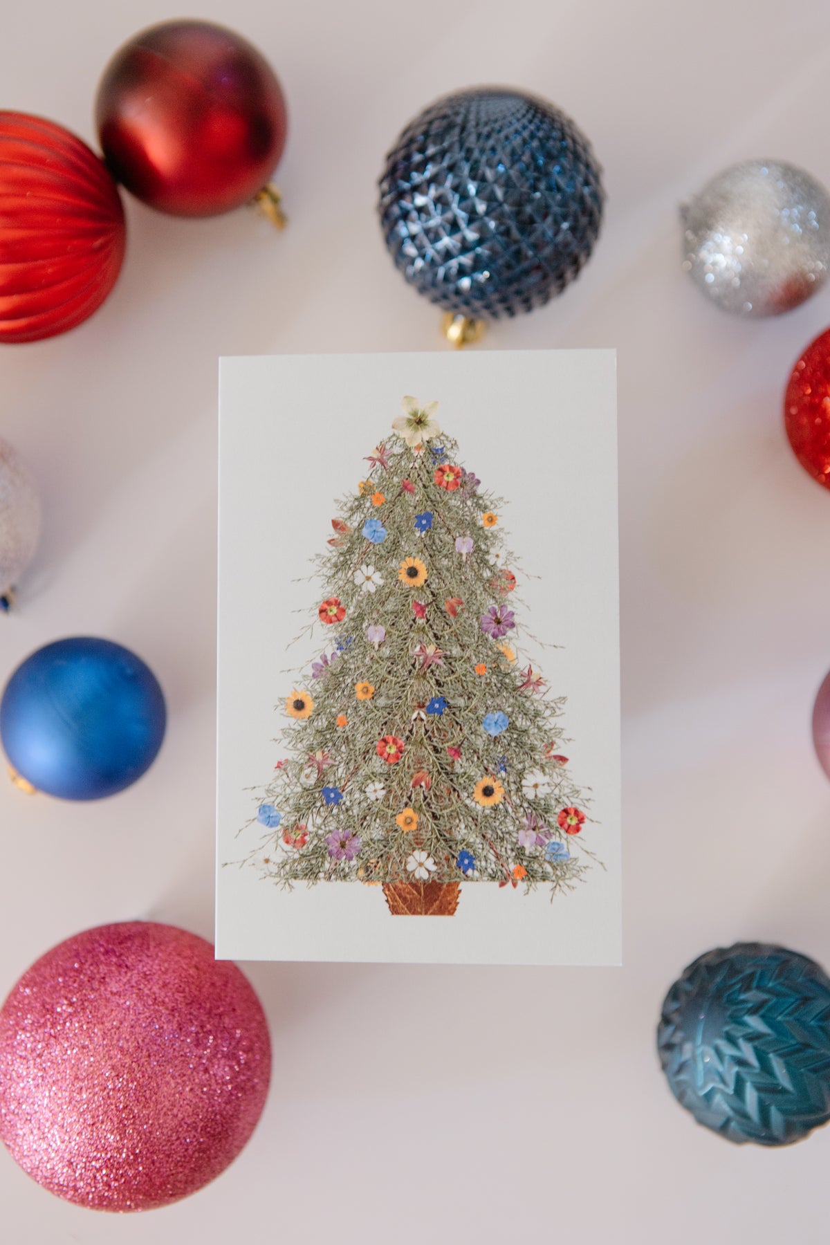 Decorative Christmas card with a floral tree design surrounded by colorful ornaments on a light background.