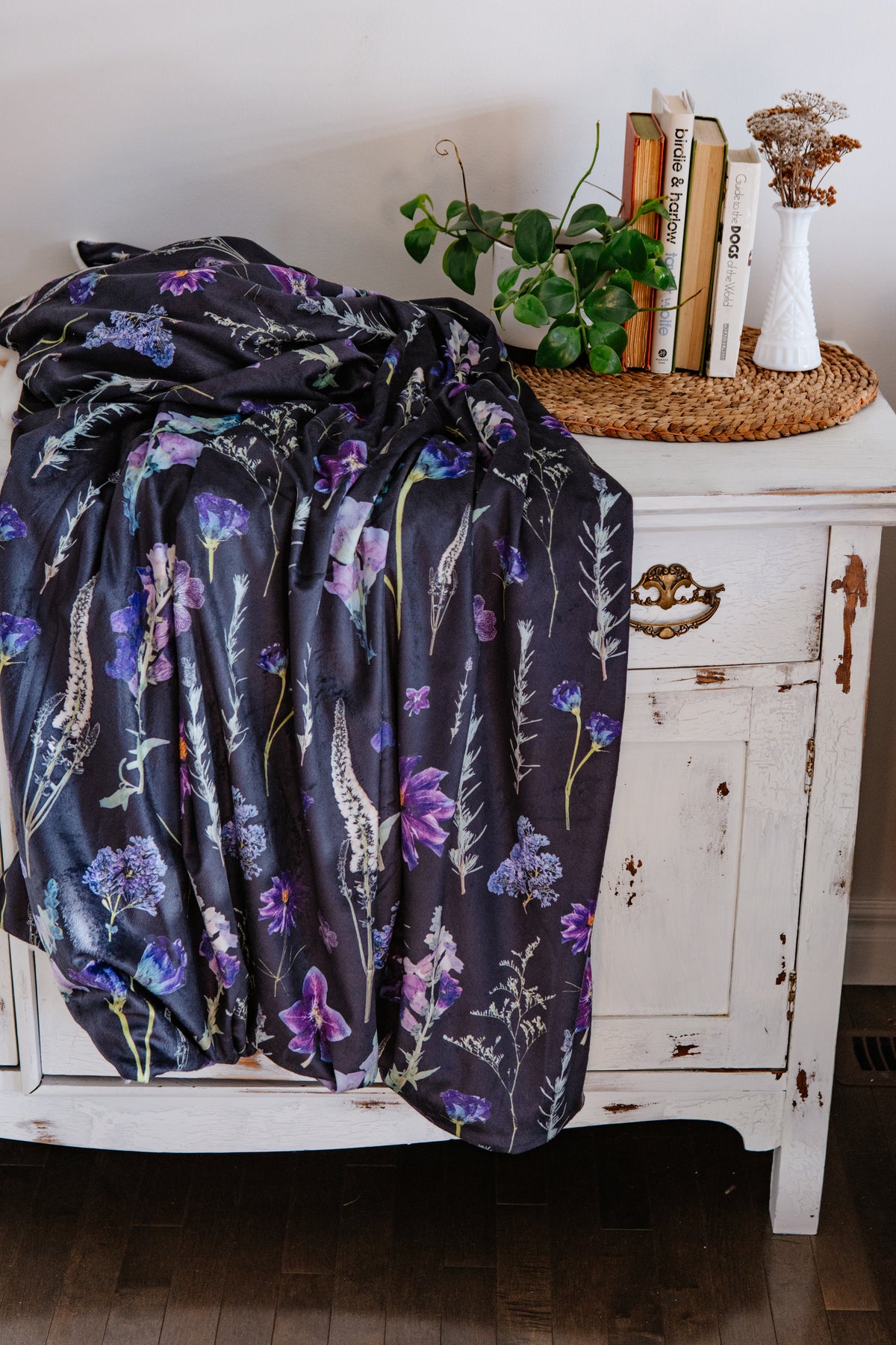 Pressed floral-patterned blanket draped over a white cabinet with books and a plant on top.