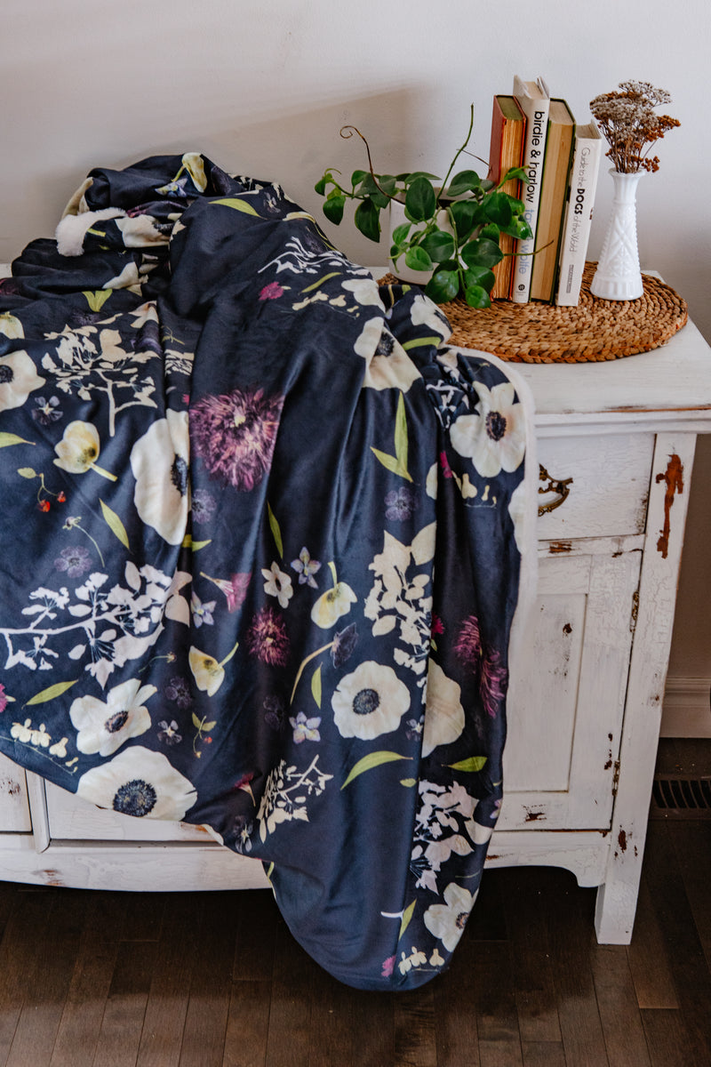 Dark navy floral blanket draped over a white cabinet with books and a plant on top.