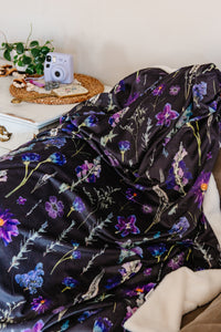 Purple floral blanket draped over a chair with a camera and plant in the background.