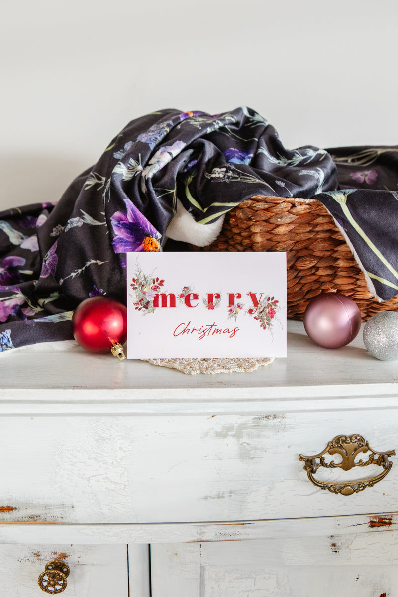 Christmas card with 'Merry Christmas' on a white surface with decorative blankets and christmas baubles.